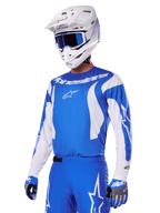 Piloto de motocross com o conjunto de equipamento Alpinestars 2025 Fluid Haul em azul e branco, camisola azul com detalhes brancos, calças azuis com logótipos de estrela, capacete branco com óculos azuis e luvas em azul e cinzento, equipamento completo de corrida todo-o-terreno