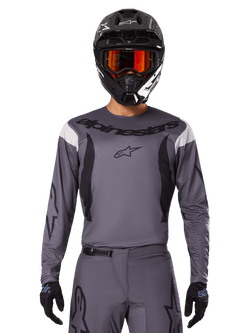 Piloto de motocross com a camisola e conjunto de equipamento Alpinestars 2025 Fluid Haul, cinzento com detalhes a preto e branco, mangas compridas e logótipos em destaque, capacete preto com óculos cor de laranja e luvas pretas, conjunto coordenado para corridas todo-o-terreno