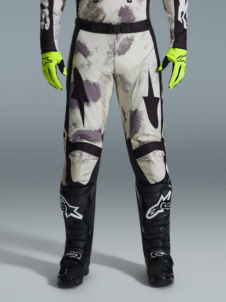 Alpinestars 2026 Racer Lahnd Pants, calças de motocross, padrão Sand Camo com grafismos abstratos em cinzento e roxo, usadas com luvas amarelo néon e botas pretas, com logótipos de estrela pretos nas coxas e cinto preto, equipamento de corrida off-road durável