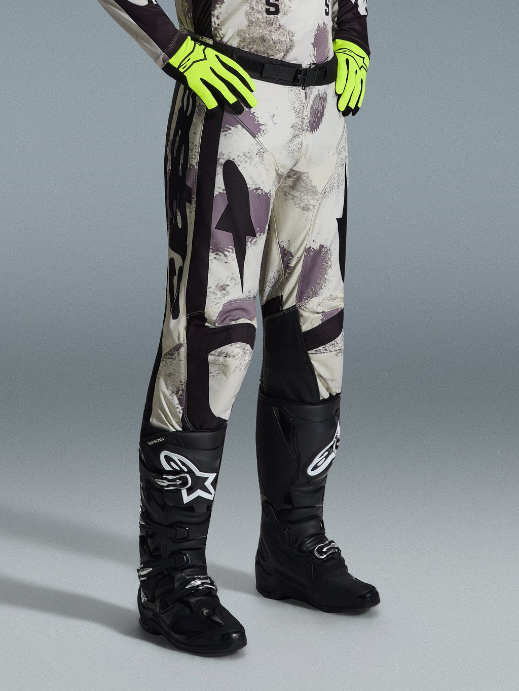 Alpinestars 2026 Racer Lahnd Pants, calças de motocross, Sand Camo, usadas com luvas amarelo néon e botas de motocross pretas, com um padrão de camuflagem abstrato, painéis laterais pretos e design ergonómico off-road