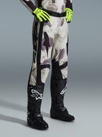 Alpinestars 2026 Racer Lahnd Pants, calças de motocross, Sand Camo, usadas com luvas amarelo néon e botas de motocross pretas, com um padrão de camuflagem abstrato, painéis laterais pretos e design ergonómico off-road