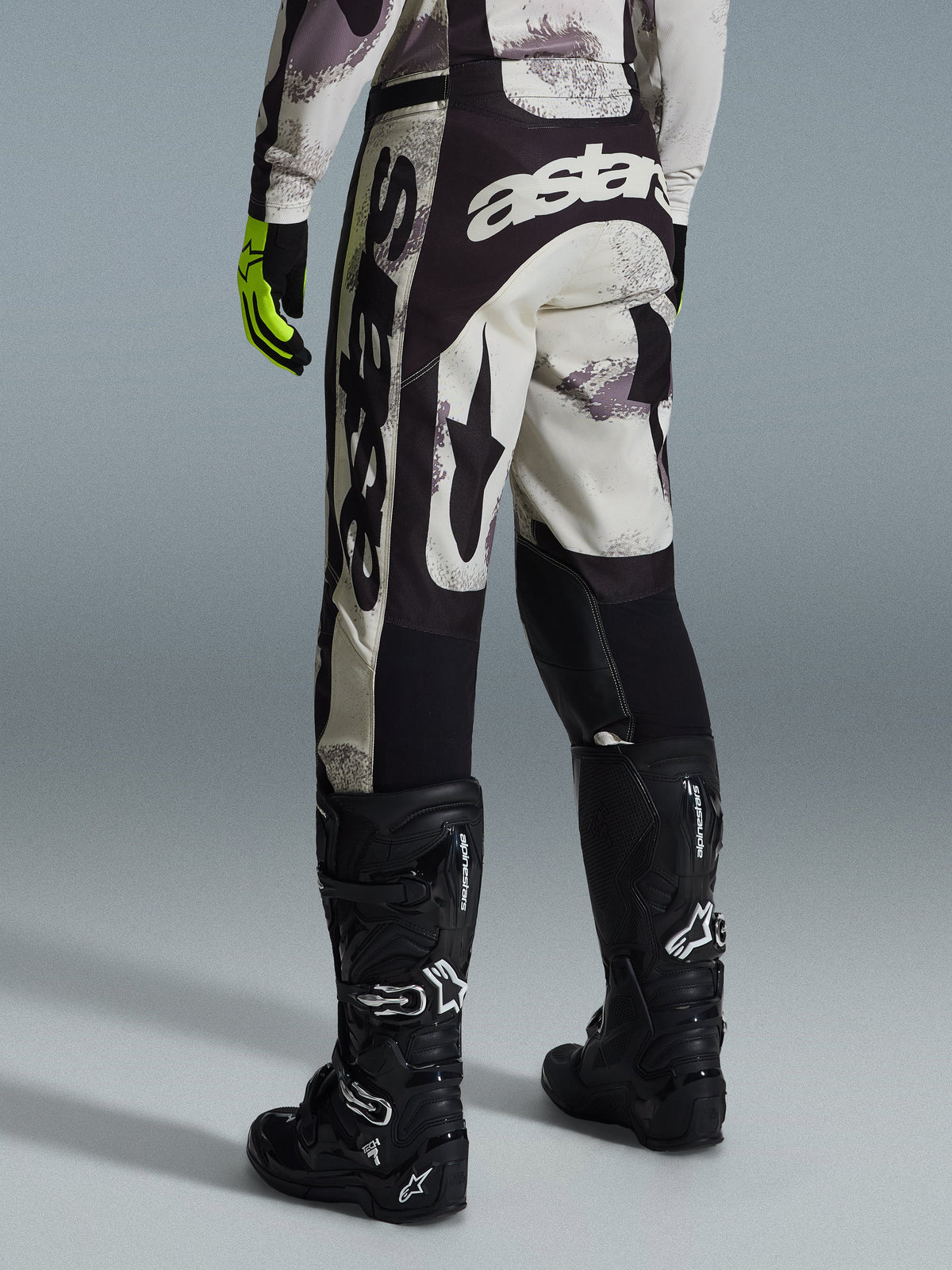 Alpinestars 2026 Racer Lahnd Pants, calças de motocross, em camuflado areia e cinzento, vista traseira apresentando logótipos brancos destacados no assento e nas pernas, combinadas com botas de motocross pretas e luvas amarelas, equipamento profissional de corrida off-road