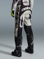 Alpinestars 2026 Racer Lahnd Pants, calças de motocross, em camuflado areia e cinzento, vista traseira apresentando logótipos brancos destacados no assento e nas pernas, combinadas com botas de motocross pretas e luvas amarelas, equipamento profissional de corrida off-road