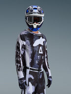 Piloto de motocross a usar a Camisola Alpinestars 2026 Racer Lahnd em Iron Camo, com um padrão de camuflagem em preto e cinzento e um grande logótipo Alpinestars branco no peito, combinada com um capacete e luvas a condizer para corridas fora de estrada.