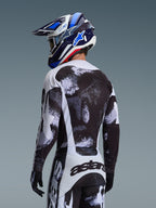Camisola Alpinestars Racer Lahnd 2026 em Iron Camo, vista traseira de um piloto de motocross com conjunto de equipamento de padrão abstrato preto, branco e cinzento, incluindo calças a condizer, luvas pretas e capacete de corrida azul com óculos de proteção.
