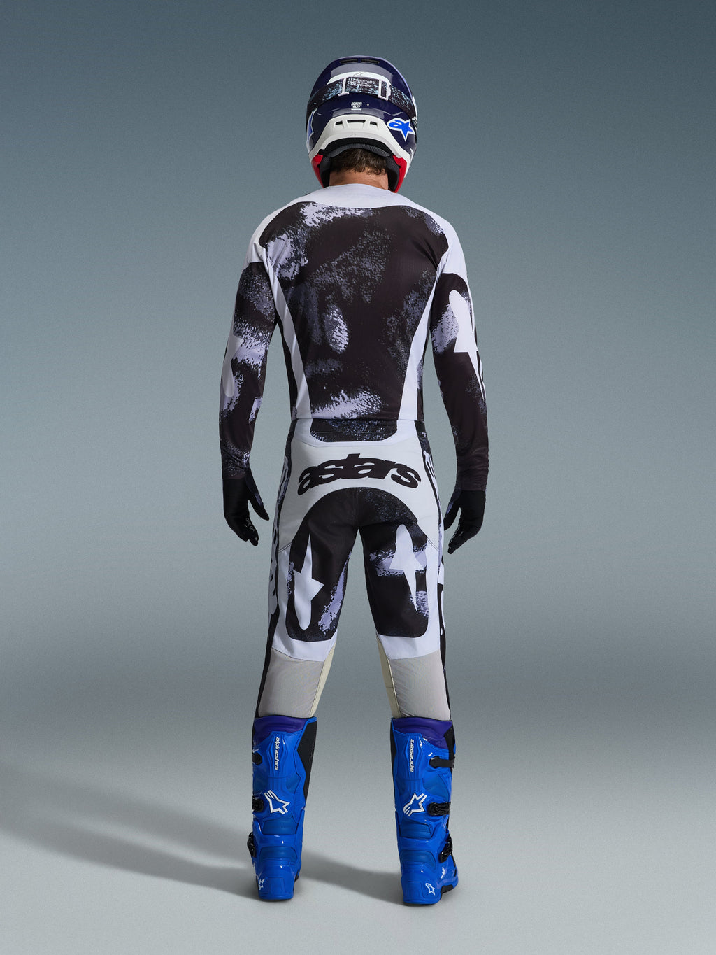 Piloto de motocross visto de costas a usar o Conjunto de Equipamento Alpinestars Racer Lahnd 2026, camisola e calças com padrão abstrato em preto e branco, logótipo 'astars' na zona do assento, combinado com botas azuis e um capacete a condizer, vestuário profissional de corrida todo-o-terreno