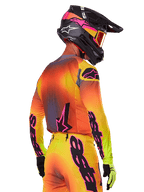 Piloto de motocross, visto de uma perspetiva lateral traseira, a usar o Conjunto de Equipamento Alpinestars Supertech Lipan 2025, com um gradiente de amarelo, laranja e vermelho, logótipos em preto e cor-de-rosa, capacete preto com detalhes rosa e óculos laranja, vestuário profissional de corrida todo-o-terreno