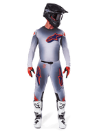 Piloto de motocross a usar o conjunto de equipamento Alpinestars Supertech Lipan 2025 em cinzento e laranja fluorescente, composto por camisola cinzenta com detalhes laranja e logótipo azul-marinho, calças cinzentas com logótipos grandes, capacete preto mate, botas brancas e pretas, e luvas vermelhas e pretas, equipamento de corrida de nível profissional.