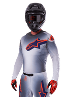 Piloto de motocross com o Conjunto de Equipamento Alpinestars Supertech Lipan 2025, camisola e calças cinzentas com detalhes em azul-marinho e laranja, capacete preto mate com óculos e luvas vermelhas e pretas, equipamento de performance atlética para corridas todo-o-terreno