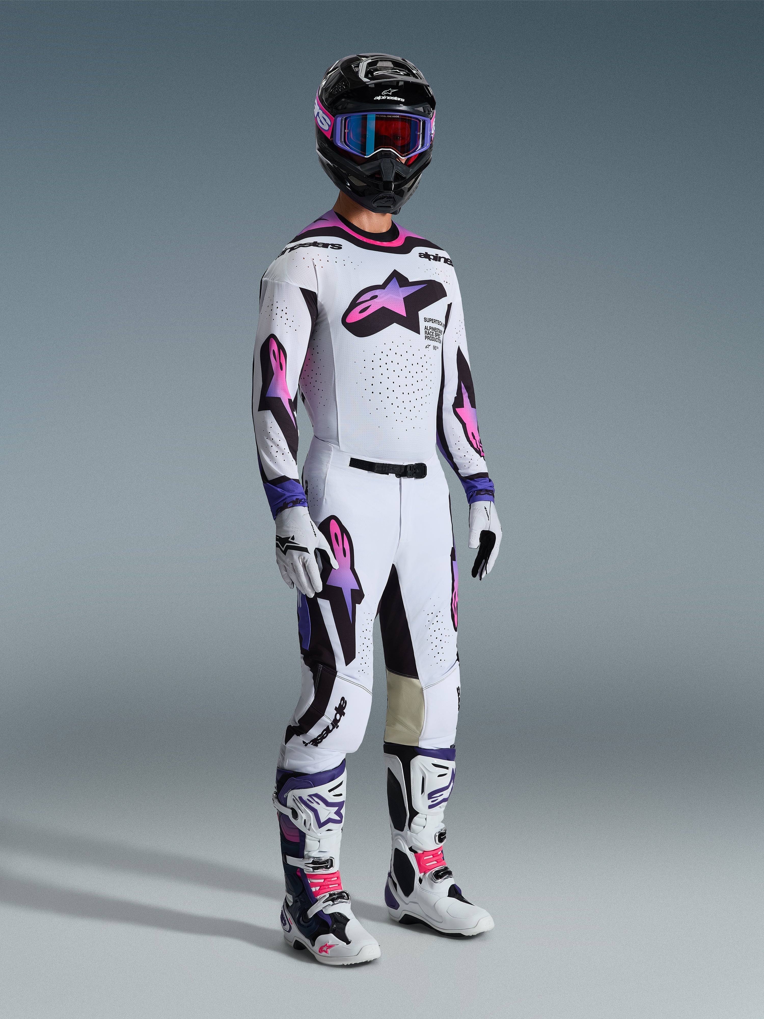 Piloto de motocross a usar a camisola Alpinestars 2026 Supertech AFD Vista em branco, roxo e preto, calças de motocross a condizer com logótipos em degradê rosa e roxo, capacete preto com óculos roxos, botas brancas e pretas, e luvas brancas, conjunto completo de equipamento profissional de corrida todo-o-terreno