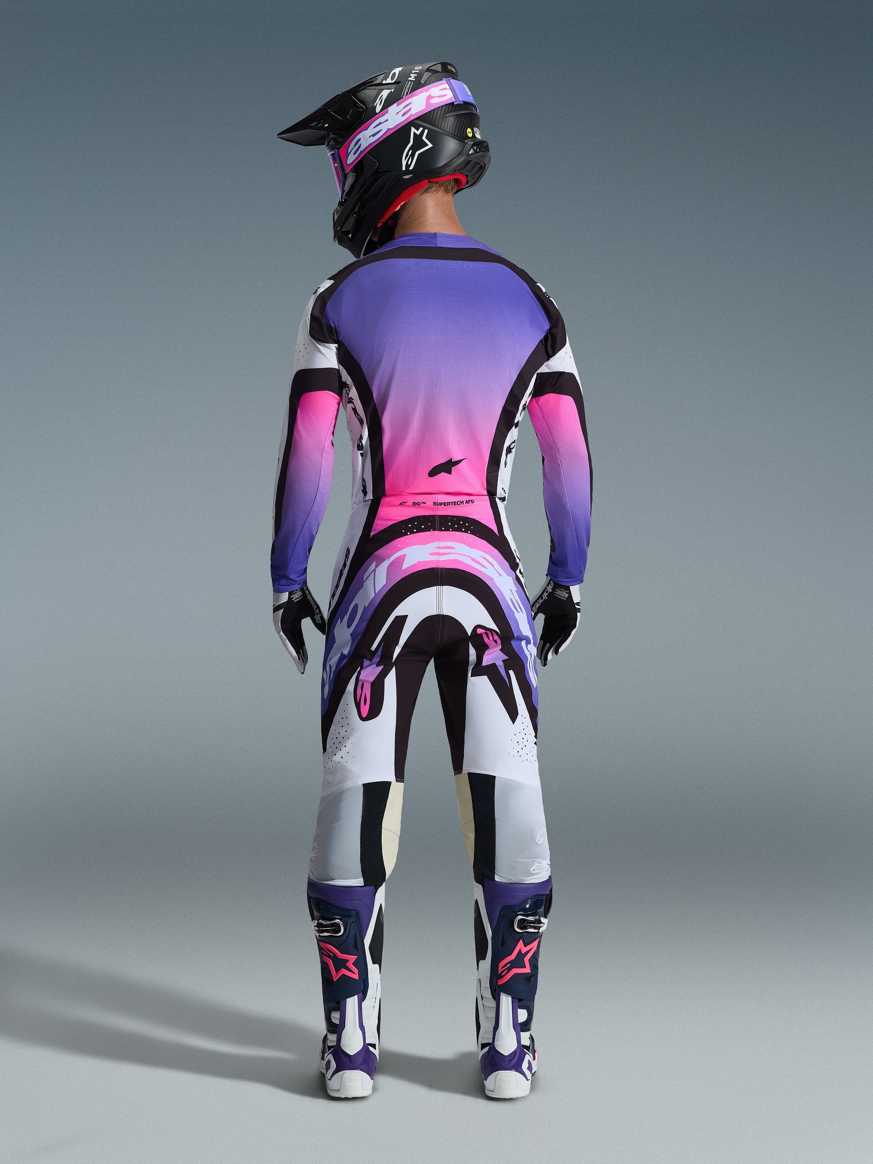 Piloto de motocross visto de costas a usar a camisola 2026 Supertech AFD Vista, branca, roxa e preta com detalhes em degradê rosa, calças de motocross a condizer, capacete preto com óculos rosa e botas brancas e roxas, conjunto completo de equipamento de corrida profissional