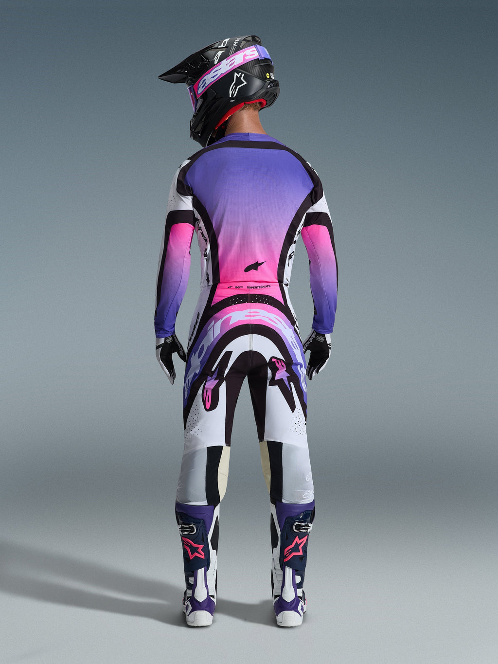 Rear view of a motocross rider wearing the Alpinestars 2026 Supertech AFD Vista Gear Set, jersey and pants featuring a vibrant white, purple, and pink gradient with black accents, black helmet with purple goggles, matching purple and white boots, coordinated professional racing outfit for off-road performance.