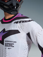 Camisola Alpinestars Supertech AFD Vista 2026, camisola de motocross, branca, roxa e preta, vista aproximada com perfurações cortadas a laser e branding race spec, usada com um capacete preto, equipamento profissional de corrida todo-o-terreno