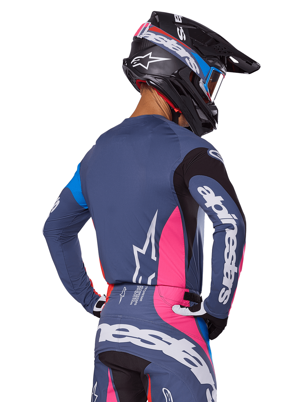 Piloto de motocross visto de costas a usar o conjunto de equipamento Alpinestars 2025 Techstar Dreem em cinzento, composto por camisola e calças azul-cinza com painéis cor-de-rosa e pretos, logótipos Alpinestars brancos e capacete preto com grafismos azuis e cor-de-rosa