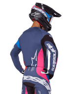 Piloto de motocross visto de costas a usar o conjunto de equipamento Alpinestars 2025 Techstar Dreem em cinzento, composto por camisola e calças azul-cinza com painéis cor-de-rosa e pretos, logótipos Alpinestars brancos e capacete preto com grafismos azuis e cor-de-rosa