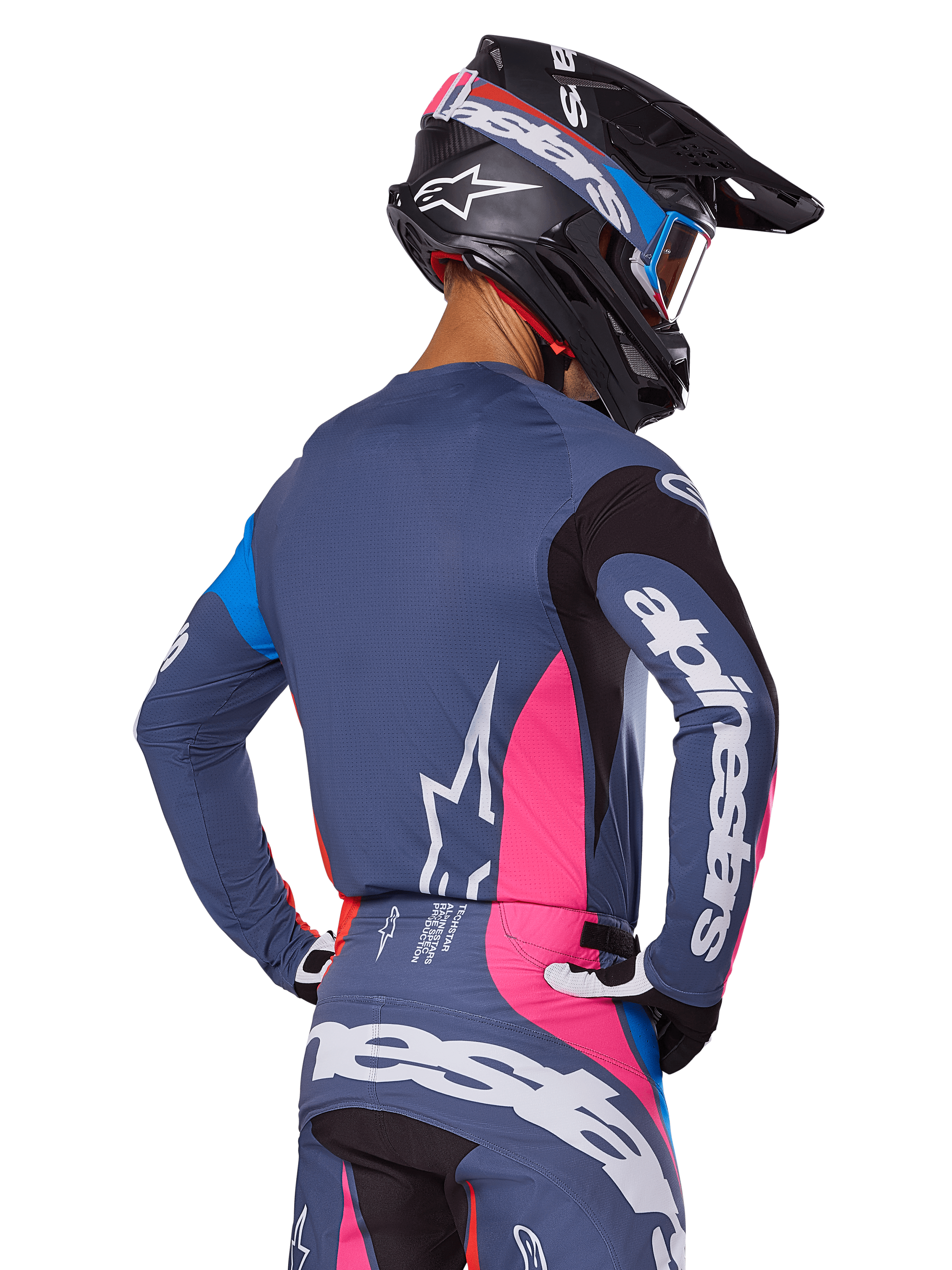 Piloto de motocross visto de costas a usar o conjunto de equipamento Alpinestars 2025 Techstar Dreem em cinzento, composto por camisola e calças azul-cinza com painéis cor-de-rosa e pretos, logótipos Alpinestars brancos e capacete preto com grafismos azuis e cor-de-rosa