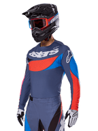 Alpinestars 2025 Techstar Dreem Gear Set, piloto de motocross com camisola e calças cinzento-ardósia com gráficos arrojados em vermelho e azul, marca 'astars' no peito, capacete preto, óculos de armação vermelha e luvas de corrida brancas