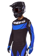 Piloto de motocross com o conjunto de equipamento Alpinestars 2025 Techstar Dreem, camisola preta com painéis azuis de estilo perfurado e logótipos brancos, calças pretas e azuis a condizer, capacete de corrida preto e luvas azuis, conjunto de performance coordenado para motocross todo-o-terreno