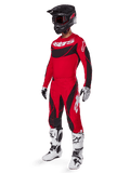 Vista de corpo inteiro de um piloto de motocross com o conjunto de equipamento Techstar Dreem 2025 em vermelho brilhante e preto, composto por camisola de performance com logótipo branco, calças ripstop elásticas a condizer, capacete preto com viseira vermelha, luvas brancas e pretas e botas Tech brancas, fato de corrida especializado para competição todo-o-terreno.