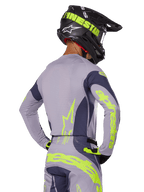 Piloto de motocross visto de costas a usar o conjunto de equipamento Alpinestars 2025 Techstar Dreem, camisola e calças cinzentas com logótipos em amarelo-verde néon vibrante, capacete preto a condizer com grafismos néon, equipamento de corrida de motocross profissional