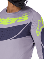 Alpinestars 2025 Techstar Dreem Jersey, camisola de motocross, cinzenta e azul-marinho com logótipo verde lima vibrante, grande plano de um piloto com capacete preto, apresentando perfurações cortadas a laser para ventilação