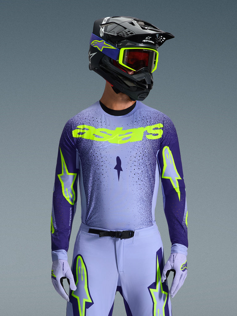 Alpinestars 2026 Supertech Scenz Jersey, camisola de motocross, roxo e amarelo fluo, vestida por um piloto com um capacete preto e roxo a condizer, óculos amarelo néon e calças de corrida roxas, com a marca astars amarelo néon no peito e logótipos de estrela nas mangas.