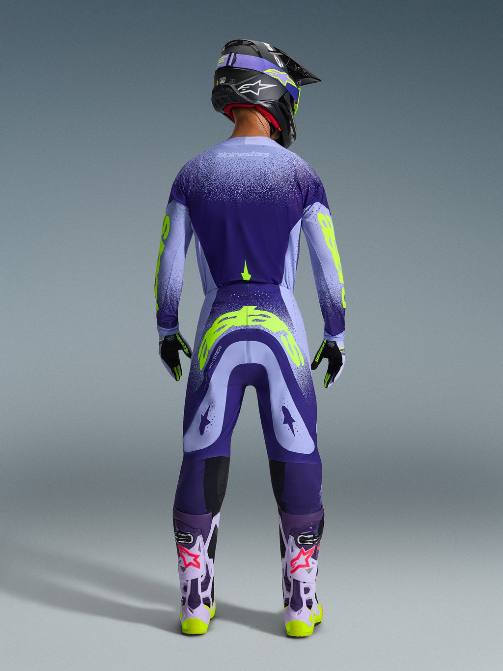 Motocross rider from behind wearing the Alpinestars 2026 Supertech Scenz Jersey in Purple Yellow Fluo, featuring a purple gradient and yellow branding, styled with matching motocross pants and purple boots, complete off-road racing kit.
