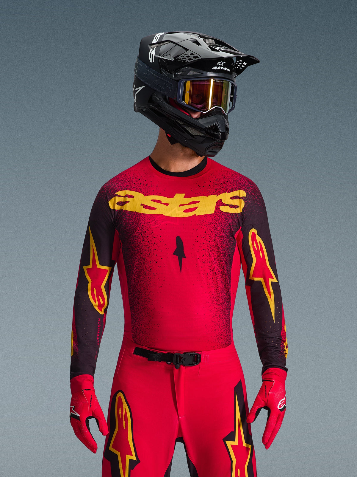 Alpinestars 2026 Supertech Scenz Jersey, camisola de motocross, vermelha, preta e amarela, usada por um piloto com um capacete preto a condizer e luvas vermelhas, com o logótipo 'astars' amarelo no peito e um padrão salpicado em degradê preto nos ombros.