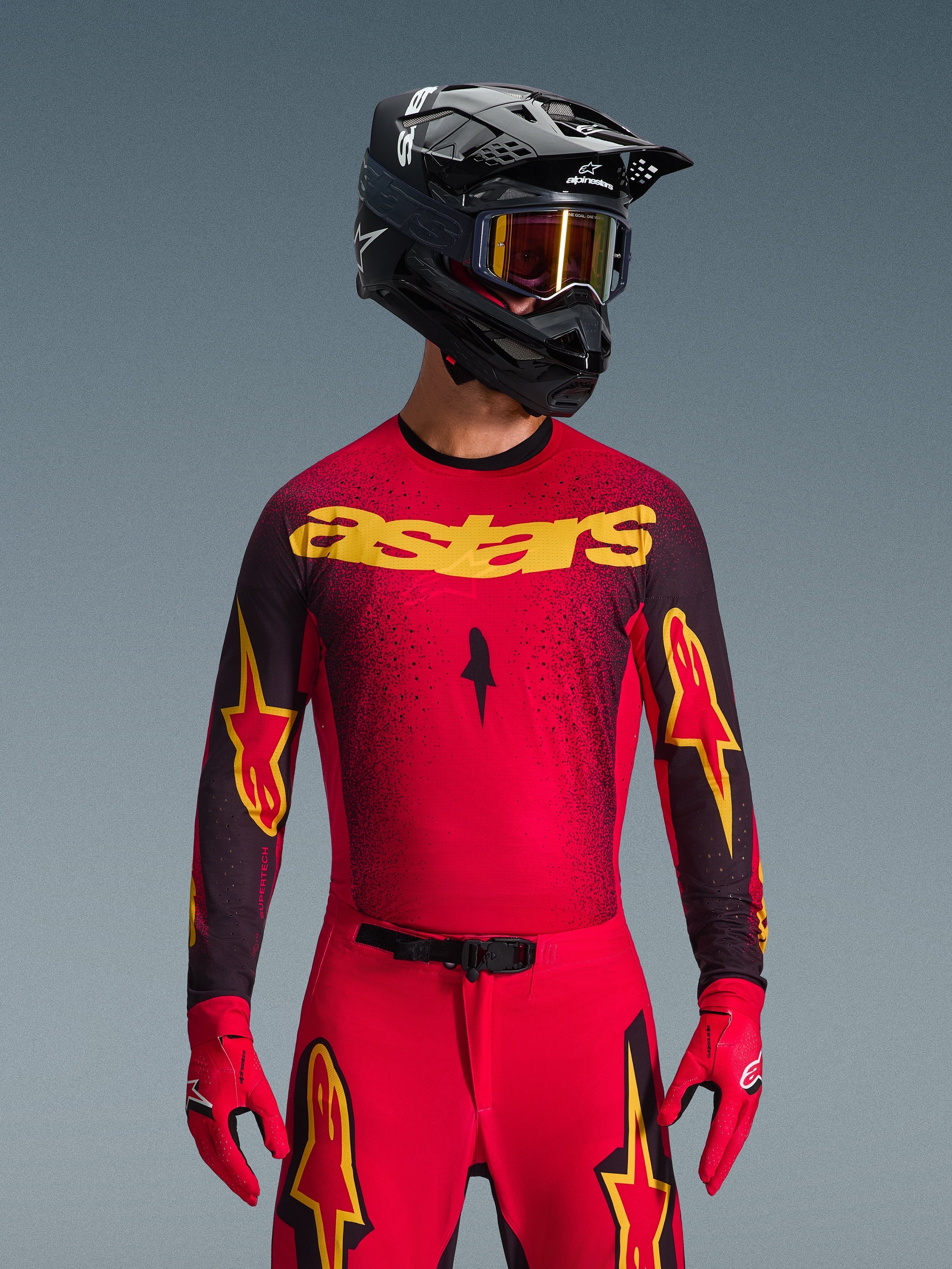 Alpinestars 2026 Supertech Scenz Jersey, camisola de motocross, vermelha, preta e amarela, usada por um piloto com um capacete preto a condizer e luvas vermelhas, com o logótipo 'astars' amarelo no peito e um padrão salpicado em degradê preto nos ombros.