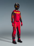 Piloto de motocross com a camisola Alpinestars Supertech Scenz 2026 em vermelho, preto e amarelo, calças vermelhas a condizer com detalhes em preto e amarelo, capacete preto com óculos, luvas vermelhas e botas pretas com detalhes vermelhos, conjunto completo de equipamento de corrida profissional