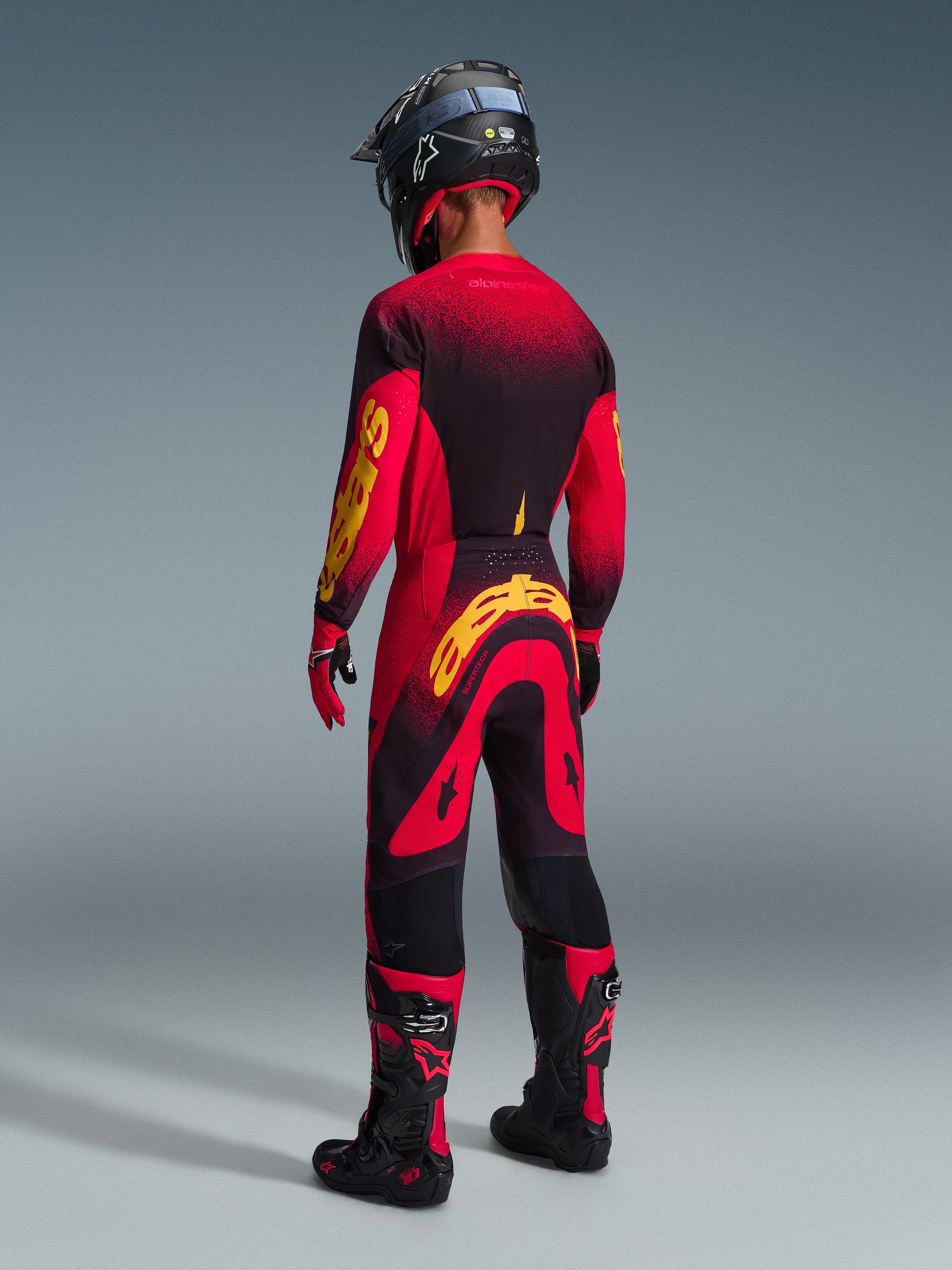 Piloto de motocross visto de costas com equipamento de corrida completo, camisola vermelha com detalhes pretos e amarelos, calças vermelhas e pretas a condizer com um logótipo Alpinestars amarelo grande, capacete preto, botas pretas e vermelhas, luvas vermelhas e pretas, conjunto coordenado para corridas profissionais de todo-o-terreno