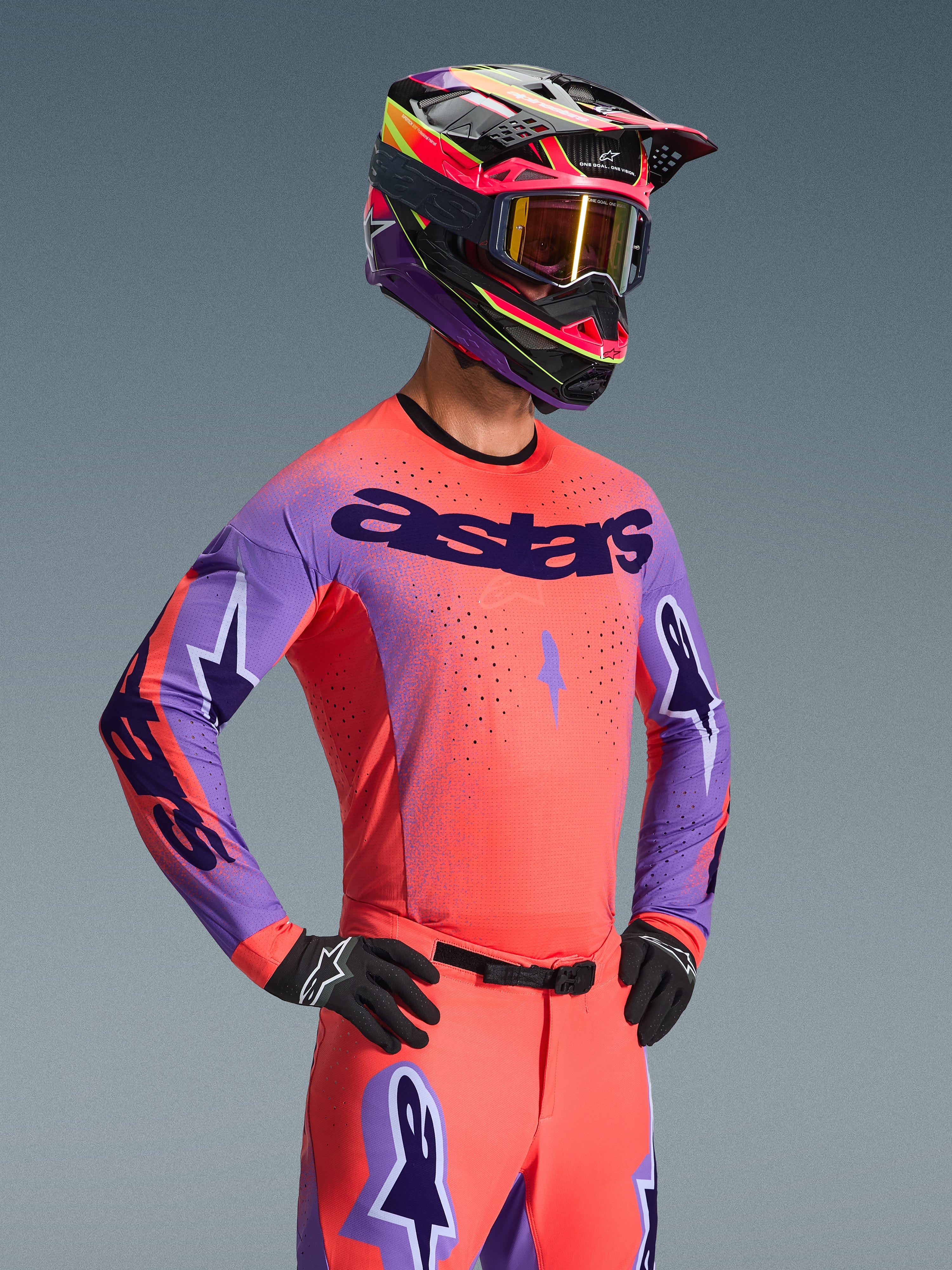 Piloto de motocross a usar a Camisola Alpinestars Supertech Scenz 2026, em Hot Coral e roxo com logótipo 'astars' de destaque no peito, equipamento a combinar incluindo calças coral e capacete coordenado em roxo e coral, com perfurações cortadas a laser para corridas todo-o-terreno de alta performance.