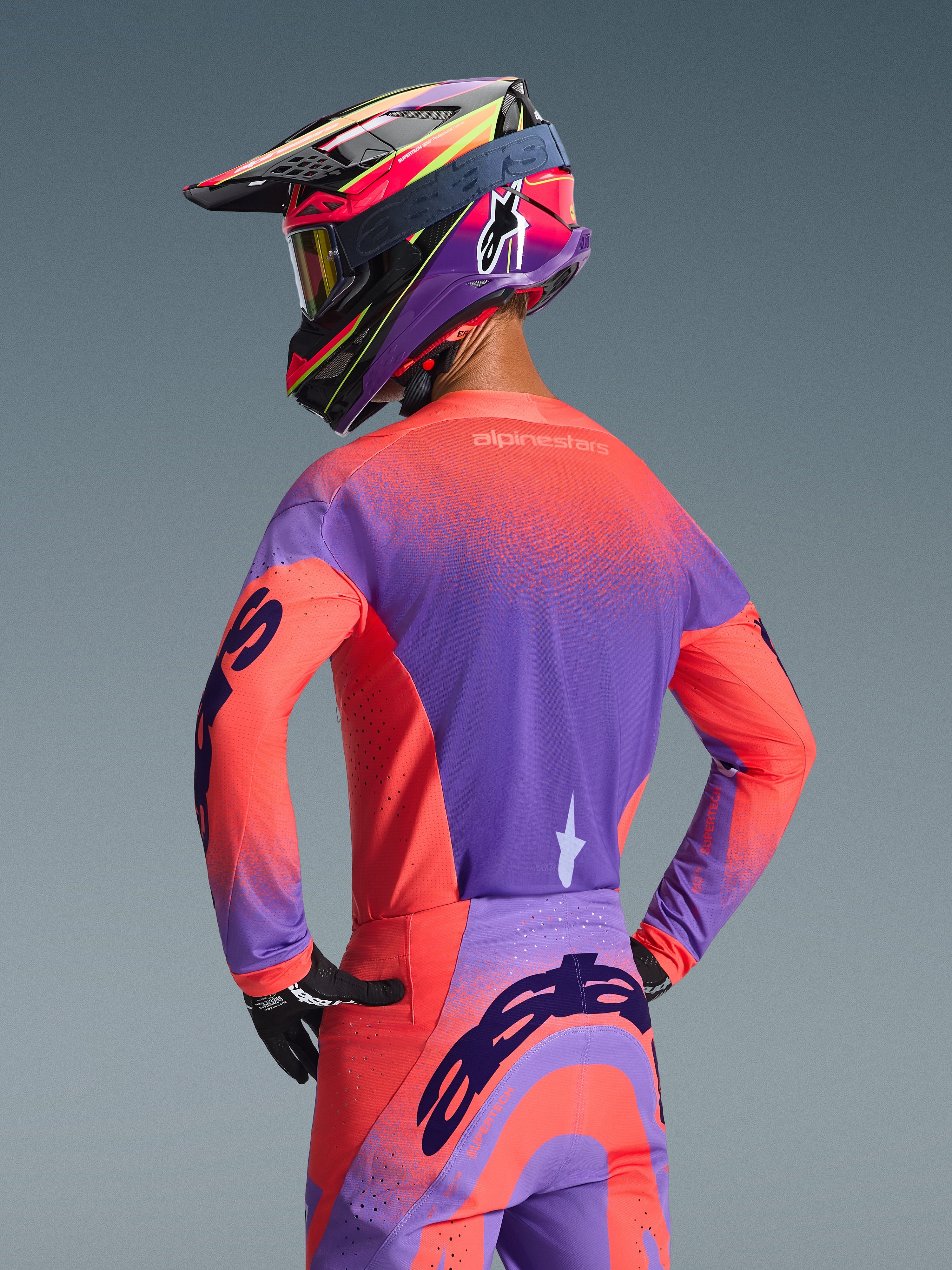 Piloto de motocross visto de trás a usar a camisola Alpinestars Supertech Scenz 2026 em Coral Vivo e Roxo, apresentando um design vibrante de gradiente salpicado, combinada com calças de motocross a condizer e um capacete de corrida colorido, equipamento profissional para desempenho todo-o-terreno.