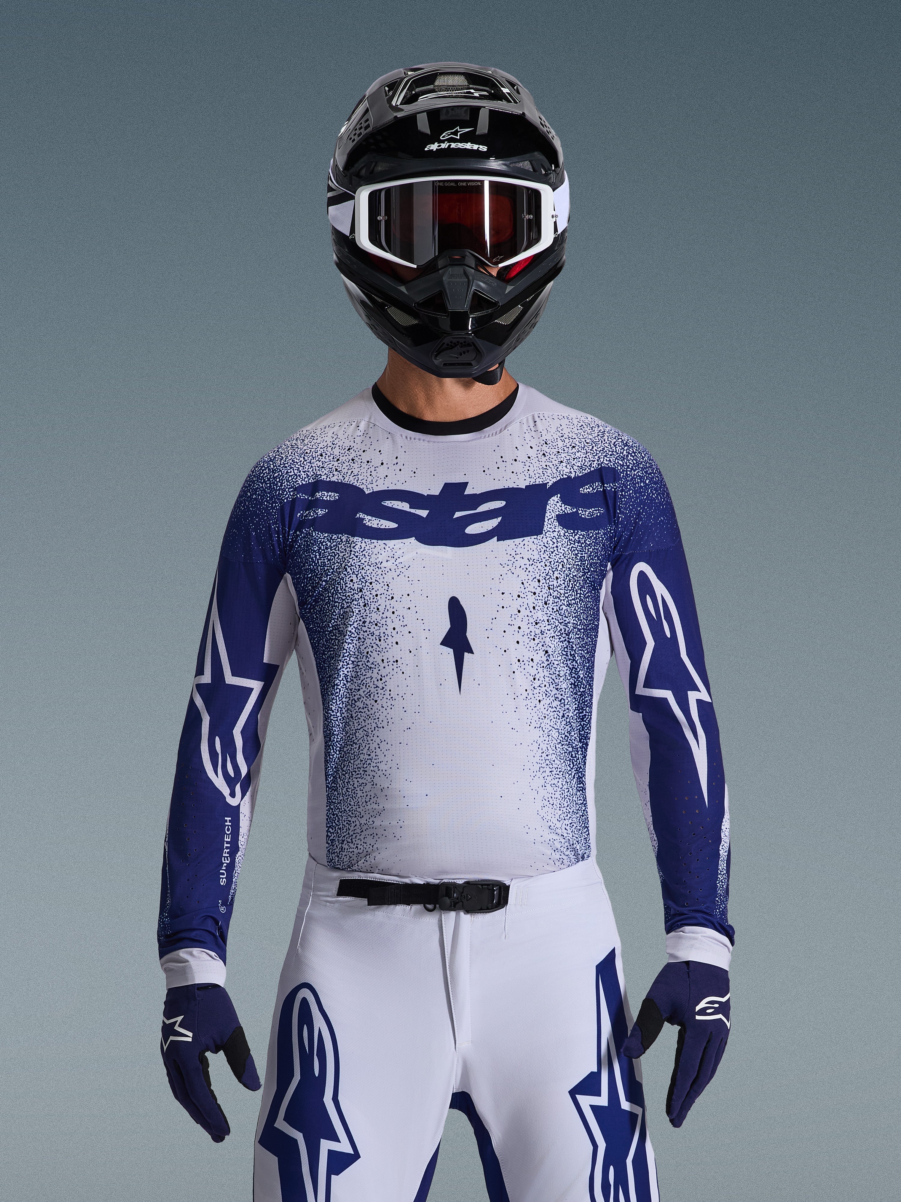 Piloto de motocross a usar a Camisola Alpinestars Supertech Scenz 2026, em cinzento claro e azul escuro com um design de gradiente salpicado e a marca 'astars' em destaque. Combinada com calças de motocross brancas a condizer, luvas azul-escuro e um capacete preto com óculos, um conjunto coordenado para competição profissional de todo-o-terreno.