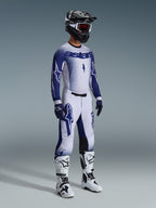 Piloto de motocross com equipamento profissional completo, camisola Supertech Scenz 2026 em cinzento claro e azul escuro, calças de motocross a combinar com grafismos de estrela, capacete preto com óculos vermelhos, botas de alta performance brancas e pretas, e luvas azuis e brancas coordenadas para competição off-road
