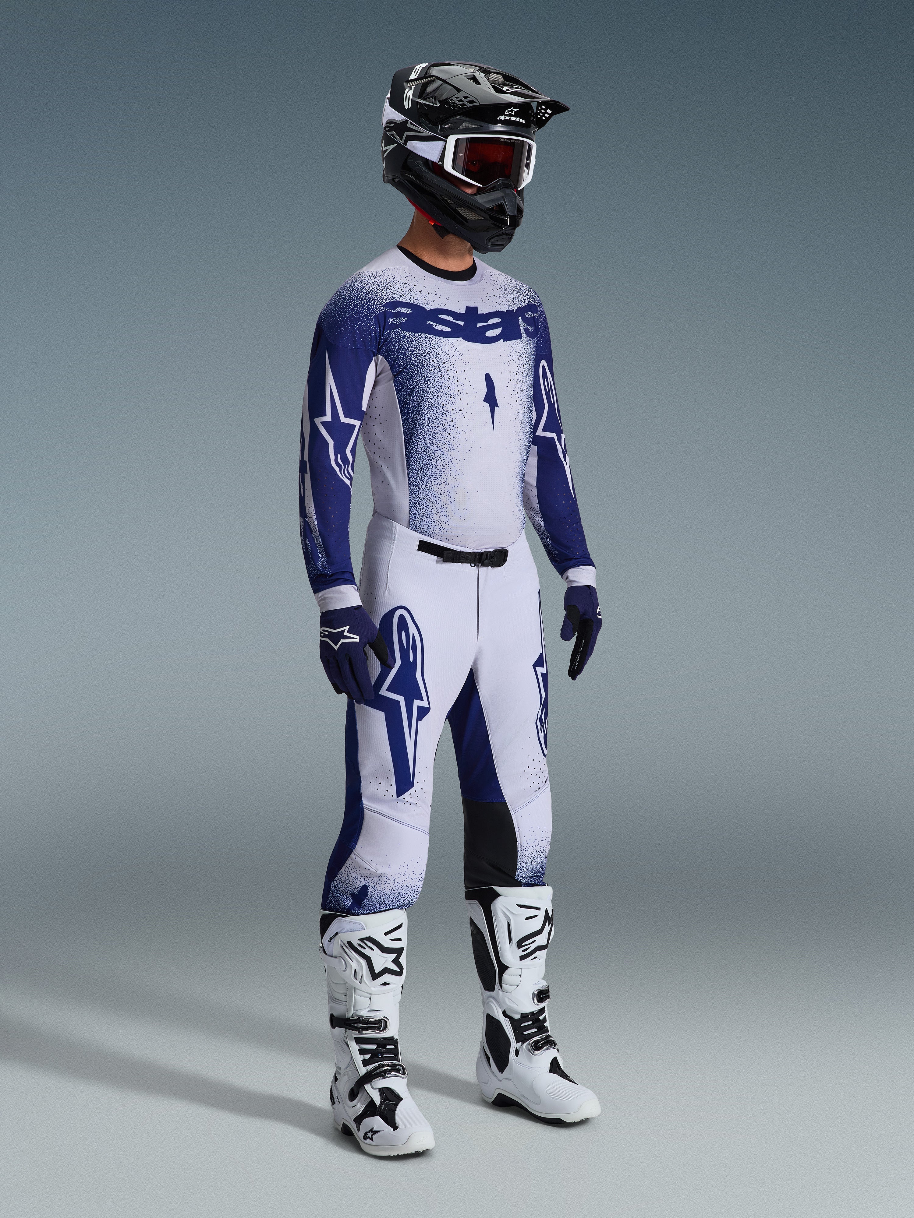 Piloto de motocross com equipamento profissional completo, camisola Supertech Scenz 2026 em cinzento claro e azul escuro, calças de motocross a combinar com grafismos de estrela, capacete preto com óculos vermelhos, botas de alta performance brancas e pretas, e luvas azuis e brancas coordenadas para competição off-road