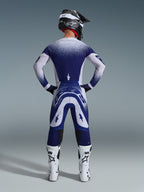 Piloto de motocross visto de trás com a camisola Alpinestars Supertech Scenz 2026, cinzento claro e azul escuro com um design de gradiente pontilhado, calças de motocross a condizer, botas de corrida brancas e capacete preto, equipamento completo de corrida todo-o-terreno.