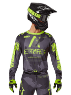 Alpinestars 2025 Racer Hollow Jersey, camisola de motocross, camuflado escuro e amarelo néon, usada com calças a condizer, capacete preto e amarelo com óculos, e luvas pretas, com a marca Alpinestars e design camuflado