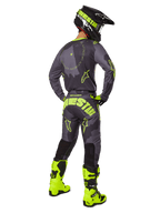 Vista traseira de um piloto de motocross a usar o conjunto de equipamento Racer Hollow 2025 em Dark Camo Yellow Fluo, com uma camisola camuflada cinzenta com detalhes em amarelo néon, calças de motocross a condizer com marca de destaque no assento, botas em amarelo néon brilhante e um capacete preto com óculos amarelos.