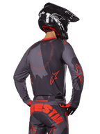 Alpinestars 2025 Racer Hollow Jersey, vista traseira de um piloto de motocross com equipamento em Cinzento Camuflado e Laranja Fluo, calças camufladas a condizer com letras Alpinestars grandes em laranja, capacete preto, design de vestuário profissional para todo-o-terreno