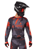 Alpinestars 2025 Racer Hollow Jersey, camisola de motocross, camuflado cinzento e laranja fluorescente, usada com conjunto de equipamento a condizer incluindo calças e luvas, com capacete preto com gráficos brancos e óculos, marca ASTARS em destaque, vestuário desportivo de proteção para motocross