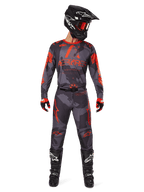 Piloto de motocross com camisola Alpinestars 2025 Racer Hollow e calças a condizer em Camo Cinzento e Laranja Fluo, conjunto completo de equipamento com capacete preto, óculos, luvas e botas, com logótipos laranja vibrantes e padrão de camuflagem.