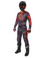 Piloto de motocross com equipamento de corrida completo, camisola e calças Alpinestars 2025 Racer Hollow em Cinzento Camuflado Laranja Fluo, capacete preto com óculos cor de laranja, luvas pretas e vermelhas, e botas pretas com logótipos brancos, conjunto de performance coordenado para corridas todo-o-terreno