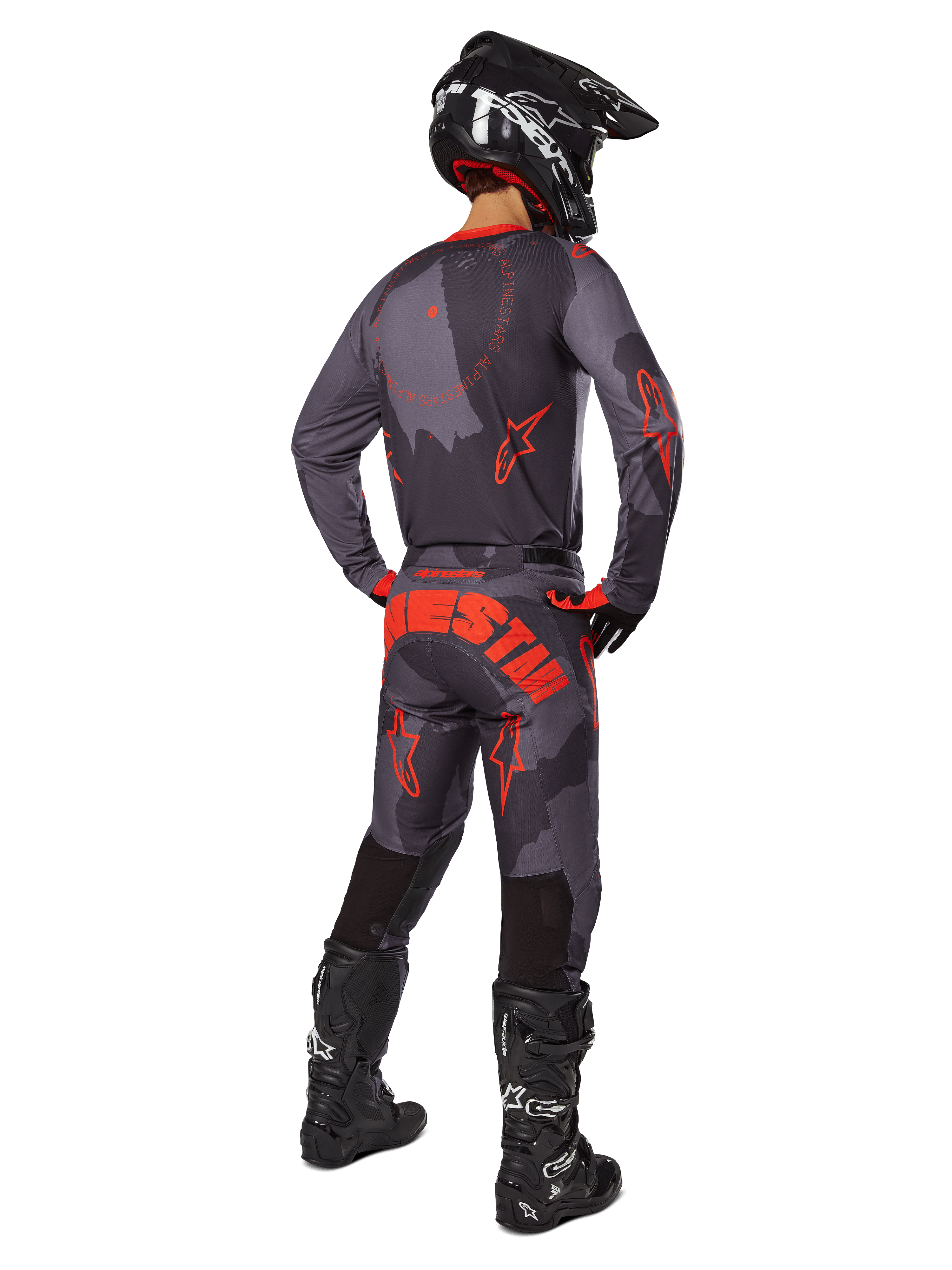 Piloto de motocross visto de trás, a usar a camisola Alpinestars 2025 Racer Hollow em Gray Camo Orange Fluo, calças camufladas a condizer com o texto Alpinestars em cor-de-laranja, capacete preto e botas pretas, conjunto de equipamento coordenado para condução todo-o-terreno