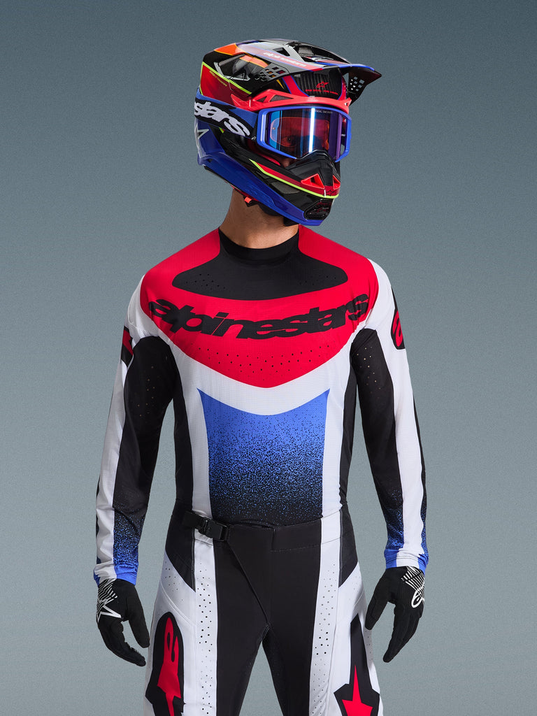 Alpinestars 2026 Techstar Knif Jersey, camisola de motocross, vermelha, branca e azul, com um logótipo marcante no peito e tronco em degradê, usada por um piloto com capacete multicolorido a condizer, óculos azuis e luvas pretas, equipamento profissional de corrida todo-o-terreno