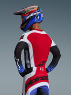 Piloto de motocross visto de costas a usar o Conjunto Techstar Knif 2026, camisola vermelha com painéis laterais brancos e mangas pretas com punhos salpicados a azul, calças vermelhas e brancas com a marca Alpinestars a preto na zona do assento, capacete coordenado azul e vermelho com óculos.