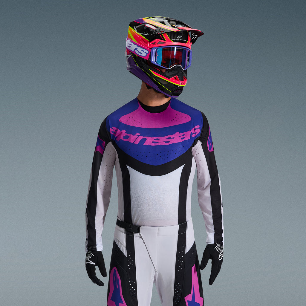 Piloto de motocross a usar o Conjunto de Equipamento Alpinestars 2026 Techstar Knif, camisola com grafismo azul e rosa no peito e tronco branco, calças brancas com painéis pretos e logótipos rosa, capacete multicolorido em roxo e rosa com óculos a condizer, luvas pretas, equipamento profissional de corrida todo-o-terreno