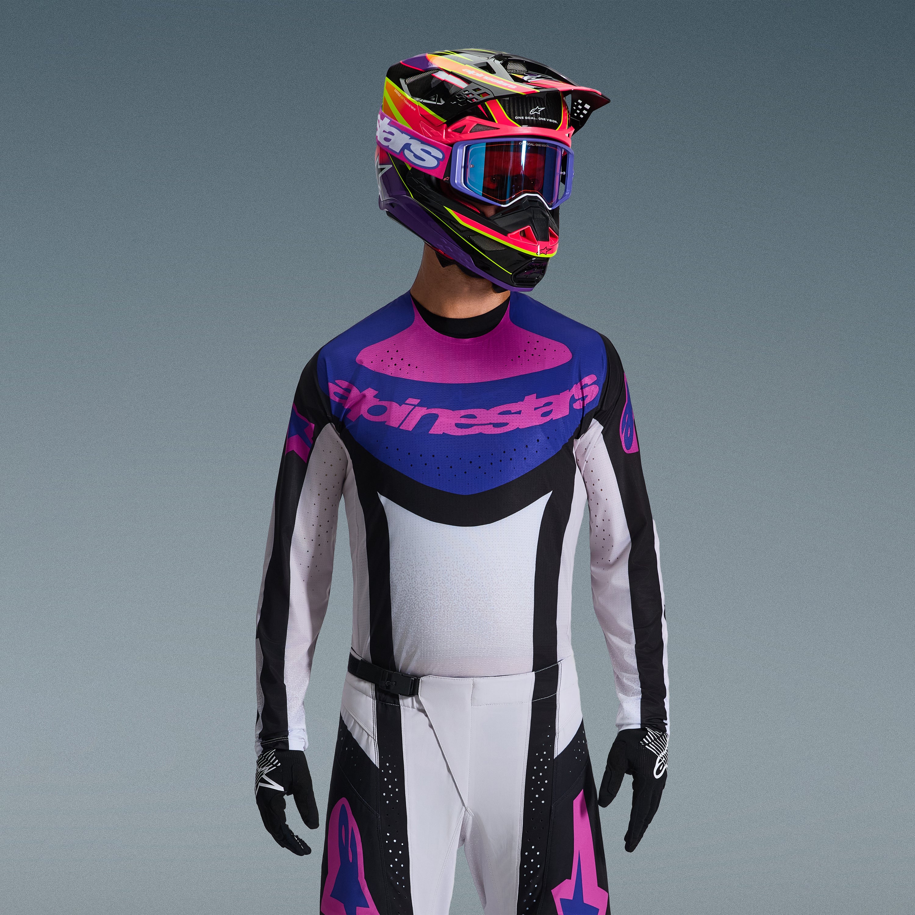 Piloto de motocross a usar o Conjunto de Equipamento Alpinestars 2026 Techstar Knif, camisola com grafismo azul e rosa no peito e tronco branco, calças brancas com painéis pretos e logótipos rosa, capacete multicolorido em roxo e rosa com óculos a condizer, luvas pretas, equipamento profissional de corrida todo-o-terreno