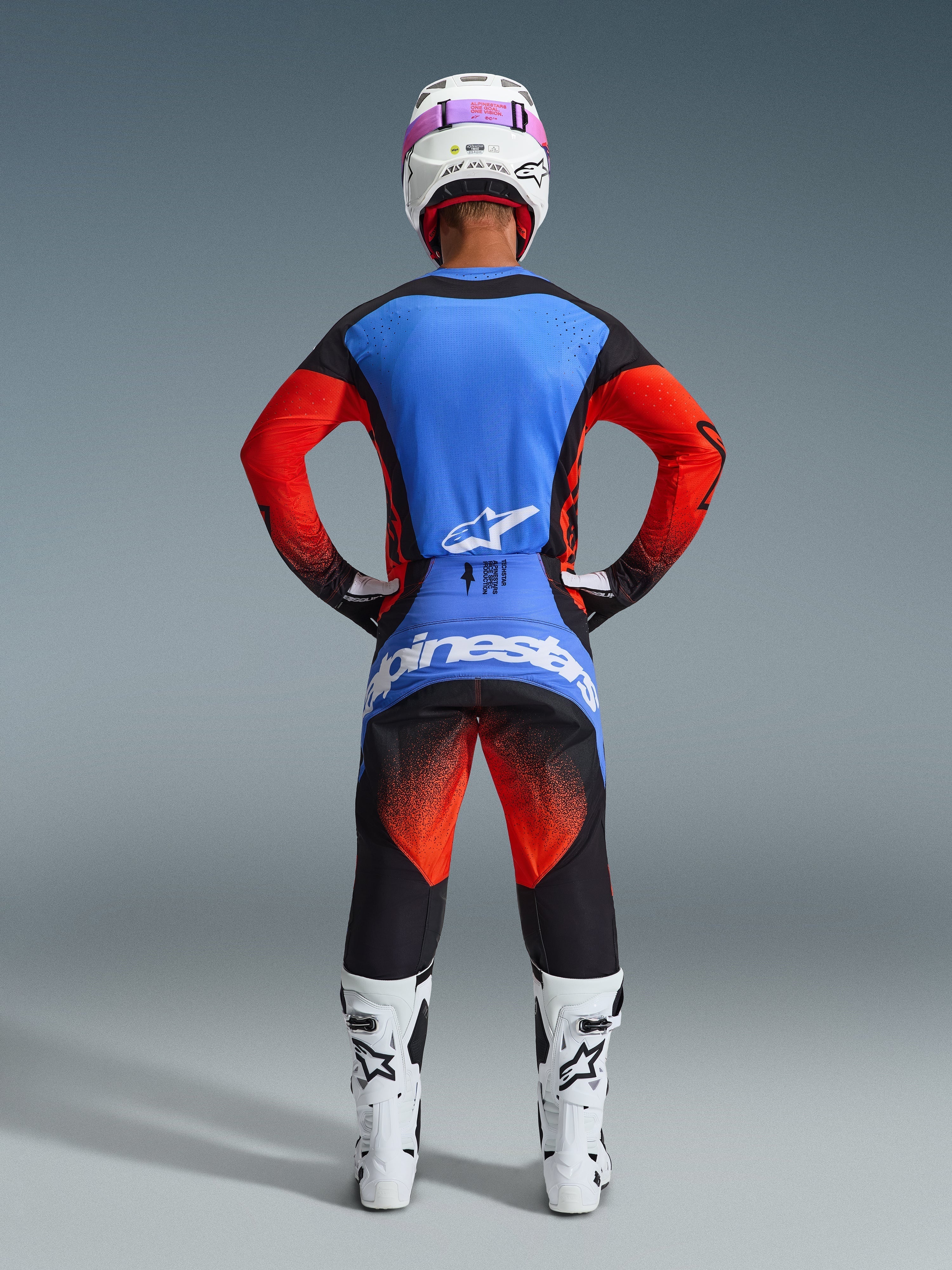 Motocross rider from behind wearing complete racing gear, 2026 Techstar Knif Jersey with blue back and orange sleeves, black pants with orange and blue accents, white helmet with pink goggle strap, white and black boots, coordinated outfit for off-road racing