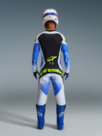 Conjunto de Equipamento Alpinestars 2026 Techstar Knif, vista traseira de um piloto de motocross em Azul Ucla, Preto e Branco, com logótipos em Amarelo Fluo na camisola e nas calças, combinado com botas de corrida azuis e capacete branco.
